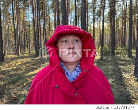 A woman stands in the forest in a red jacket and hood, thoughtfully looking at the surroundings, walking in the fresh air A woman stands in the forest in a red jacket and hood, thoughtfully looking at the surroundings, walking in the fresh air 138731377