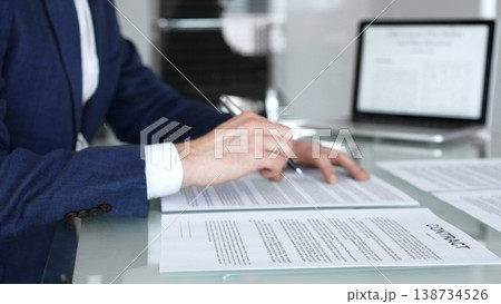 Close-up view of a business person in a professional office setting, reviewing and signing a legal contract document. Hands holding a pen over paperwork during corporate deal negotiations 138734526