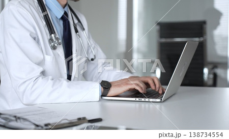 Confident doctor wearing a white coat and stethoscope, using a laptop at a desk for medical record keeping and online patient care. Medicine, healthcare and science concept Confident doctor wearing a white coat and stethoscope, using a laptop at a desk for medical record keeping and online patient care. Medicine, healthcare and science concept 138734554