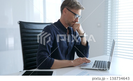 Businessman sitting at a sleek desk, working on a laptop and thinking in a bright, modern office filled with natural light. Business people concept 138734643