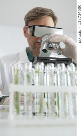 Man scientific researcher in white protective gear analyzing specimens through a microscope in a sterile laboratory environment. Medicine, health care and science concept 138734680
