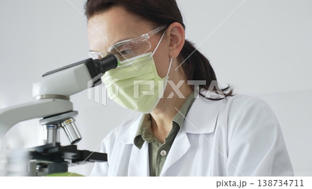 Female professional scientist examining specimen, placing slide under microscope while wearing green protective gear in laboratory workspace. Medicine, health care and science concept Female professional scientist examining specimen, placing slide under microscope while wearing green protective gear in laboratory workspace. Medicine, health care and science concept 138734711