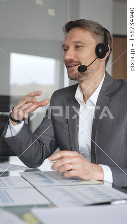 Businessman professional wearing a headset, engaging in an online video call or teleconference, discussing financial data and charts while working in a modern office setup, vertical view 138734940