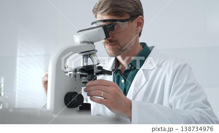 Adult male scientist wearing lab coat and protective glasses using microscope, doing scientific research in modern laboratory. Medicine, healthcare and science concept Adult male scientist wearing lab coat and protective glasses using microscope, doing scientific research in modern laboratory. Medicine, healthcare and science concept 138734976