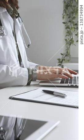 Doctor woman wearing white lab coat over green shirt, and stethoscope, is typing on laptop, documenting patient information. Medicine and health care concept Doctor woman wearing white lab coat over green shirt, and stethoscope, is typing on laptop, documenting patient information. Medicine and health care concept 138734994