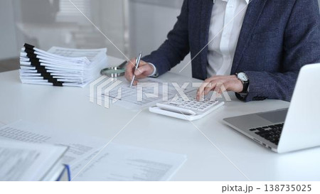 Business professional reviewing financial reports, calculating with a calculator, managing document stacks and laptop at white desk. Audit and taxes Business professional reviewing financial reports, calculating with a calculator, managing document stacks and laptop at white desk. Audit and taxes 138735025