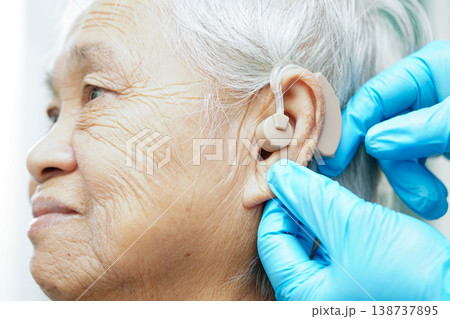 Doctor install hearing aid on senior patient ear to reduce hearing loss problem. Doctor install hearing aid on senior patient ear to reduce hearing loss problem. 138737895