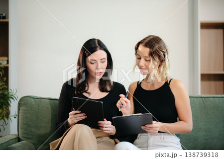 Two women collaborating on a project at home, brainstorming with a laptop and notebook in a cozy setting. Two women collaborating on a project at home, brainstorming with a laptop and notebook in a cozy setting. 138739318