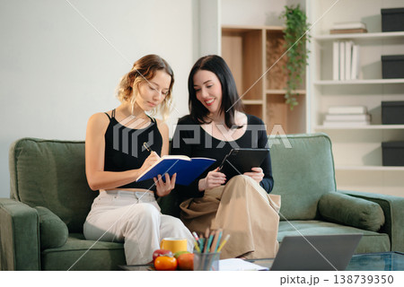 Two women collaborating on a project at home, brainstorming with a laptop and notebook in a cozy setting. Two women collaborating on a project at home, brainstorming with a laptop and notebook in a cozy setting. 138739350