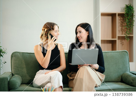 Two women collaborating on a project at home, brainstorming with a laptop and notebook in a cozy setting. Two women collaborating on a project at home, brainstorming with a laptop and notebook in a cozy setting. 138739386