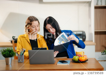 Two professional women collaborating on a business project with a laptop, notebook, and coffee at home. 138739589