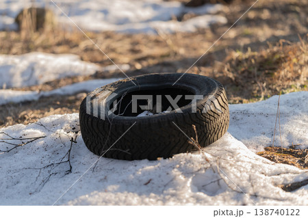 Abandoned tire scene. Vintage rubber on melting snow. Weathered wheel placed near thawing forest edge. Forgotten rubber tire amid melting snow and lush surroundings 138740122