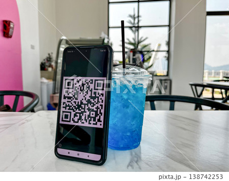 Smartphone displaying a QR code for digital payment placed on a table next to a blue drink in a cafe. Cashless transaction, mobile banking, and contactless technology in modern lifestyle setting Smartphone displaying a QR code for digital payment placed on a table next to a blue drink in a cafe. Cashless transaction, mobile banking, and contactless technology in modern lifestyle setting 138742253