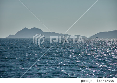 A sailboat sails at sunset through the Vigo Estuary, with the silhouettes of the Cies Islands outlined on the horizon and the shimmering waters. A sailboat sails at sunset through the Vigo Estuary, with the silhouettes of the Cies Islands outlined on the horizon and the shimmering waters. 138742550