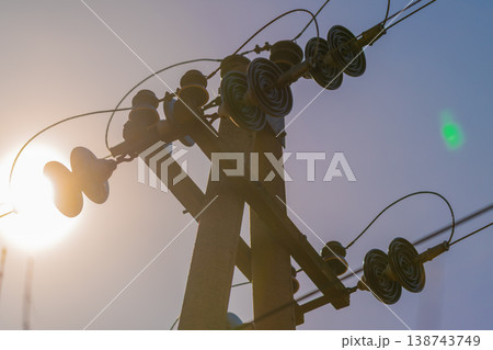 Backlit pole silhouette with electric lines, low sun creating bright flare and contrasty outlines of insulators, warm highlights and atmospheric mood photography 138743749