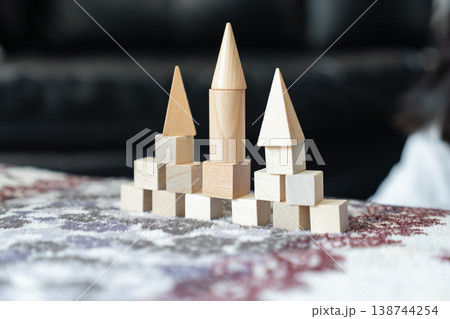 Natural wood blocks form small castle with three pointed towers as basic geometric pieces stand in ordered rows on soft fabric creating calm indoor play scene for spatial learning. 138744254