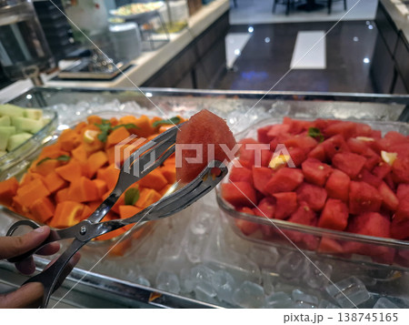 Candid Close-up of Hand Using Tongs to Pick Fresh Watermelon from Buffet Display 138745165
