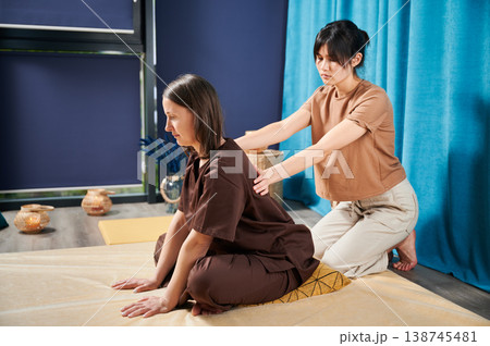 Masseuse making thai yoga massage. Woman therapist doing traditional massage treatment for female patient indoors. Client sitting on mat, enjoying hand massage in calm atmosphere. Masseuse making thai yoga massage. Woman therapist doing traditional massage treatment for female patient indoors. Client sitting on mat, enjoying hand massage in calm atmosphere. 138745481