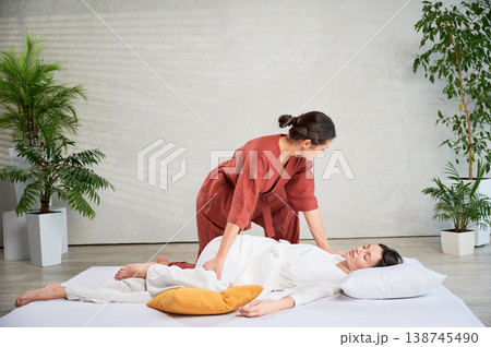 Masseuse making thai yoga massage. Woman therapist doing traditional massage treatment, stretching female patient's body. Client lying on mat, enjoying hand massage in calm atmosphere. 138745490