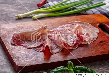 Thinly sliced cured meat, possibly prosciutto, on a wooden cutting board with green onions and basil. Thinly sliced cured meat, possibly prosciutto, on a wooden cutting board with green onions and basil. 138751403