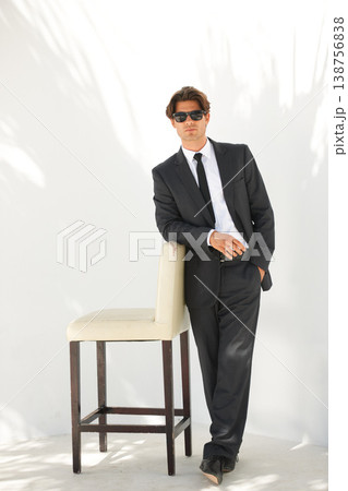 Portrait, fashion and a business man with a chair in studio on a white background for corporate style. Handsome, sunglasses and formal with a confident young professional employee leaning on a seat 138756838
