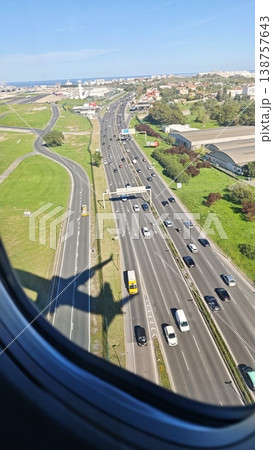 Aerial view of a busy highway with vehicles traveling, green fields and buildings visible, captured from an airplane window during descent near a coastal city Aerial view of a busy highway with vehicles traveling, green fields and buildings visible, captured from an airplane window during descent near a coastal city 138757643