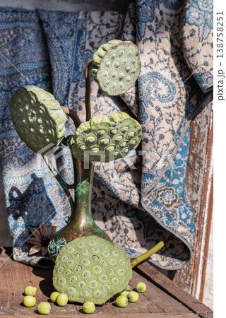 Several green lotus seed pods rise from a slender bronze-colored vase, their unique, honeycomb textures catching the light. Below, a single large pod and a handful of round seeds are strewn across a 138758251