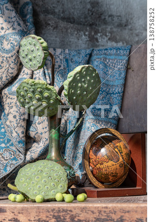 A weathered wooden chair serves as the base for a still-life featuring an ornate bronze vase and two dried lotus pods. A patterned fabric and a small wooden globe accent the arrangement, all set 138758252