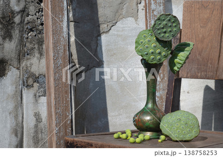 Green lotus pods are arranged in a slender bronze vase atop a weathered wooden seat. The scene is defined by rustic textures, from the peeling paint of the chair to the seeds scattered near the vase. Green lotus pods are arranged in a slender bronze vase atop a weathered wooden seat. The scene is defined by rustic textures, from the peeling paint of the chair to the seeds scattered near the vase. 138758253