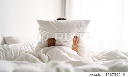Person lies in bed hiding face behind pillow in bright bedroom for privacy and comfort Person lies in bed hiding face behind pillow in bright bedroom for privacy and comfort 138759946