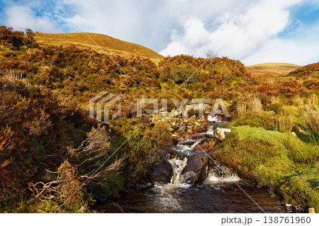 A vibrant stream tumbles over rocks and cascades down the hillside. Lush green moss and brown heather cover the hillsides beneath a bright blue sky. 138761960