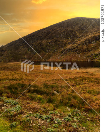 A vast mountain rises under a vibrant orange sunset sky. A calm, dark lake rests at its base, surrounded by golden and green moorland vegetation in the foreground. 138761975