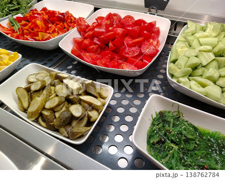 Freshly prepared vegetable salad ingredients displayed in white bowls, including red bell peppers, tomatoes, cucumbers, pickles, and seaweed on a stainless steel counter Freshly prepared vegetable salad ingredients displayed in white bowls, including red bell peppers, tomatoes, cucumbers, pickles, and seaweed on a stainless steel counter 138762784