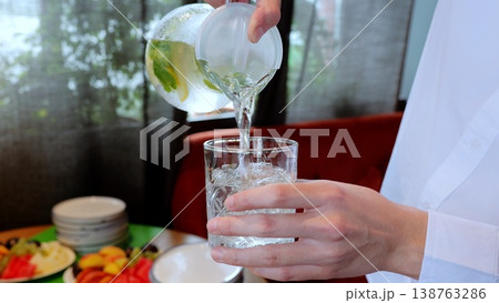 Waiter pouring chilled water with lemon and mint into glass, fruit platter decorating restaurant background 138763286