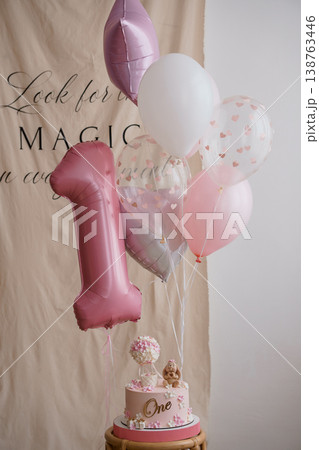 Elegant pink first birthday cake with number one balloon and floral decorations on wooden stand against neutral backdrop. 138763446