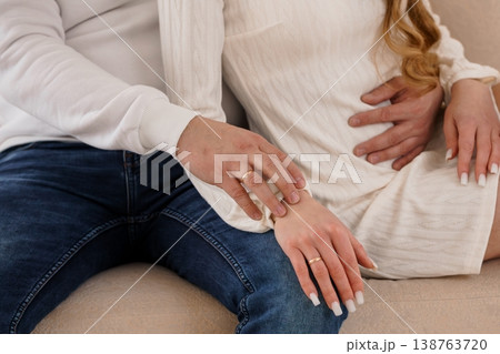 Pregnant woman holding belly with partner hand on arm, white cardigan, blue jeans, intimate couple moment, soft lighting, neutral tones. 138763720