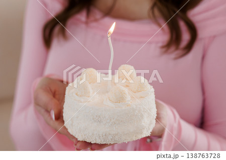 Young woman holding small white birthday cake with single lit candle and pink clothing closeup Young woman holding small white birthday cake with single lit candle and pink clothing closeup 138763728