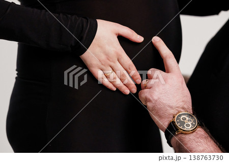 Pregnant couple holding baby bump with wedding rings and luxury watch on black background 138763730
