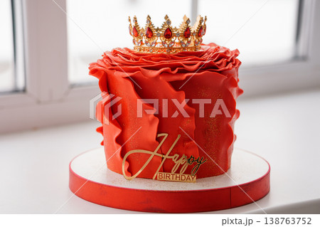Luxurious red birthday cake with gold crown topper and ruffled frosting on white window sill with natural light. Luxurious red birthday cake with gold crown topper and ruffled frosting on white window sill with natural light. 138763752