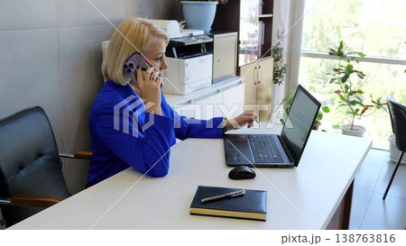 Woman in a blue suit multitasks at her desk in a modern office, working on a laptop and talking on her phone Woman in a blue suit multitasks at her desk in a modern office, working on a laptop and talking on her phone 138763816