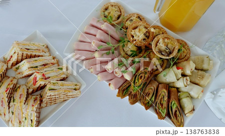 Catering service displaying gourmet finger foods ham cheese rolls, club sandwiches, mini quiches, stuffed pancakes, fresh juice arranged on crisp white linen Catering service displaying gourmet finger foods ham cheese rolls, club sandwiches, mini quiches, stuffed pancakes, fresh juice arranged on crisp white linen 138763838