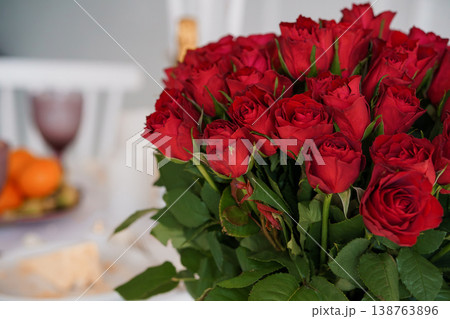 Large bouquet of vibrant red roses with green leaves on a blurred dinner table setting with wine glass and fruit in background. Large bouquet of vibrant red roses with green leaves on a blurred dinner table setting with wine glass and fruit in background. 138763896