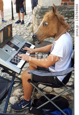 Street performer wearing a humorous horse head mask while playing an electronic keyboard on a cobblestone street 138764668