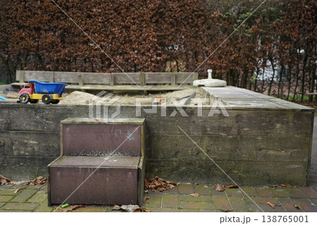 Wooden sandbox with a blue toy dump truck and a bench on a leaf-covered patio beside a low wall and trees. Quiet playground scene. Wooden sandbox with a blue toy dump truck and a bench on a leaf-covered patio beside a low wall and trees. Quiet playground scene. 138765001