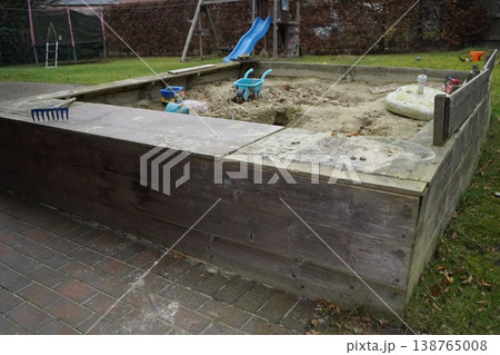 Wooden sandbox in a yard with sand, toy wheelbarrow, rake, and blue slide in the background. Outdoor play area for children. 138765008