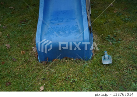 Blue plastic playground slide end on grassy lawn with a small metal anchor beside it. Outdoor play equipment shown close up. 138765014