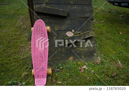 Pink plastic skateboard standing on grass beside a dark wooden box with loose planks. Outdoor yard scene with scattered leaves; a casual moment in nature. Pink plastic skateboard standing on grass beside a dark wooden box with loose planks. Outdoor yard scene with scattered leaves; a casual moment in nature. 138765020