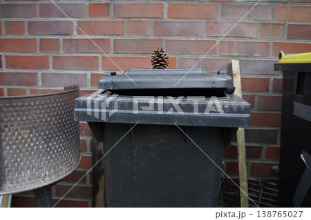 A dark plastic trash bin with an open lid stands against a brick wall, with a pine cone on top and tools nearby. Everyday outdoor waste area scene. A dark plastic trash bin with an open lid stands against a brick wall, with a pine cone on top and tools nearby. Everyday outdoor waste area scene. 138765027
