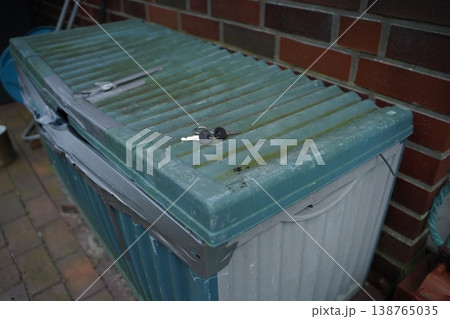 Green plastic bin lid with duct tape and a key on top, beside a brick wall on a tiled floor. It shows a taped container in a storage area. Green plastic bin lid with duct tape and a key on top, beside a brick wall on a tiled floor. It shows a taped container in a storage area. 138765035