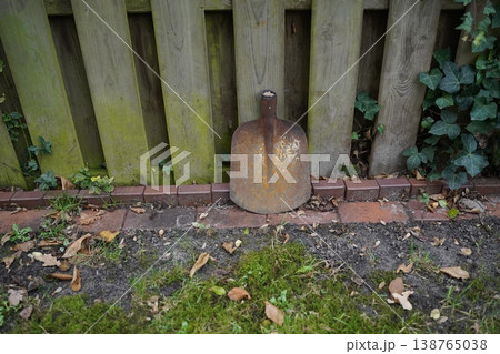 Rusted metal shovel head leaning against a wooden fence beside bricks, dirt, and grass; ivy and fallen leaves around. Weathered garden scene. Rusted metal shovel head leaning against a wooden fence beside bricks, dirt, and grass; ivy and fallen leaves around. Weathered garden scene. 138765038